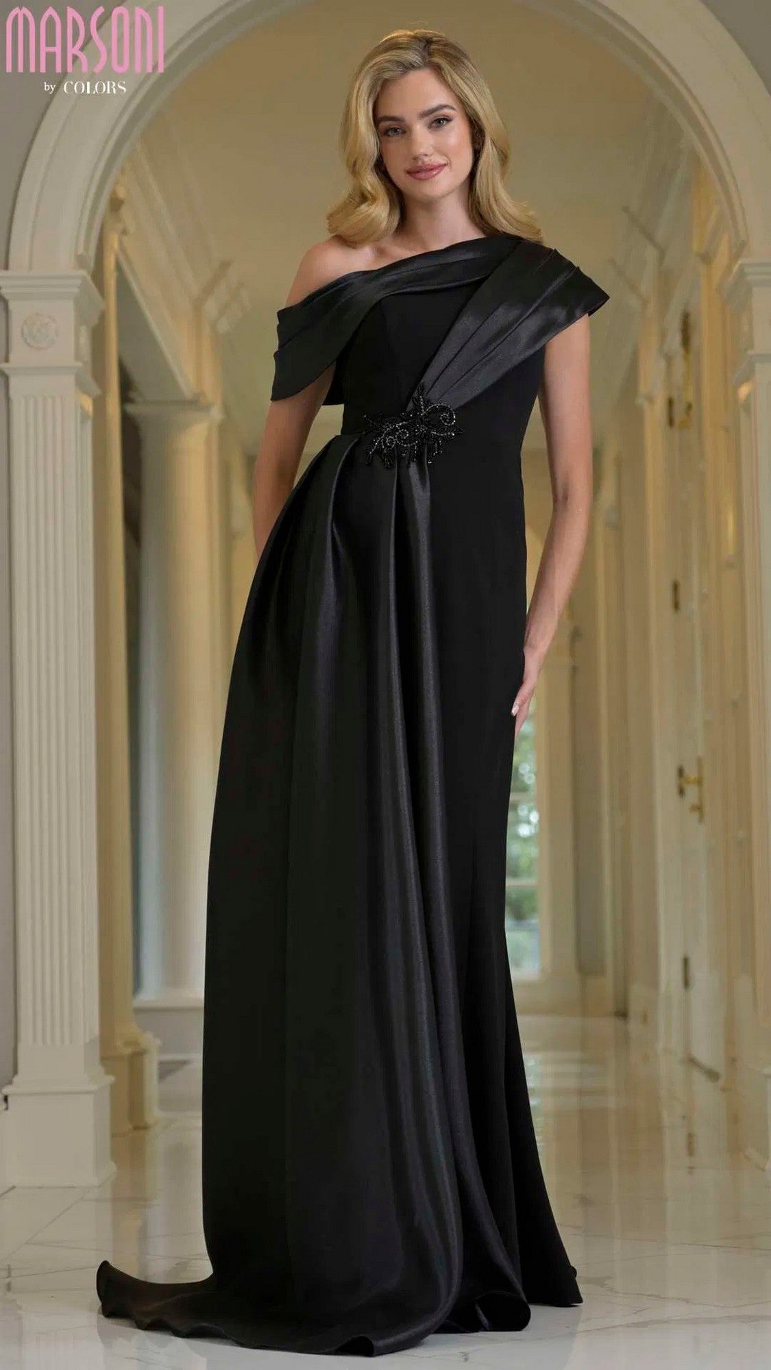 A woman stands in a hallway wearing the Marsoni M346 long formal dress in Black Black, an off-the-shoulder stretch crepe gown featuring a brooch detail at the waist.